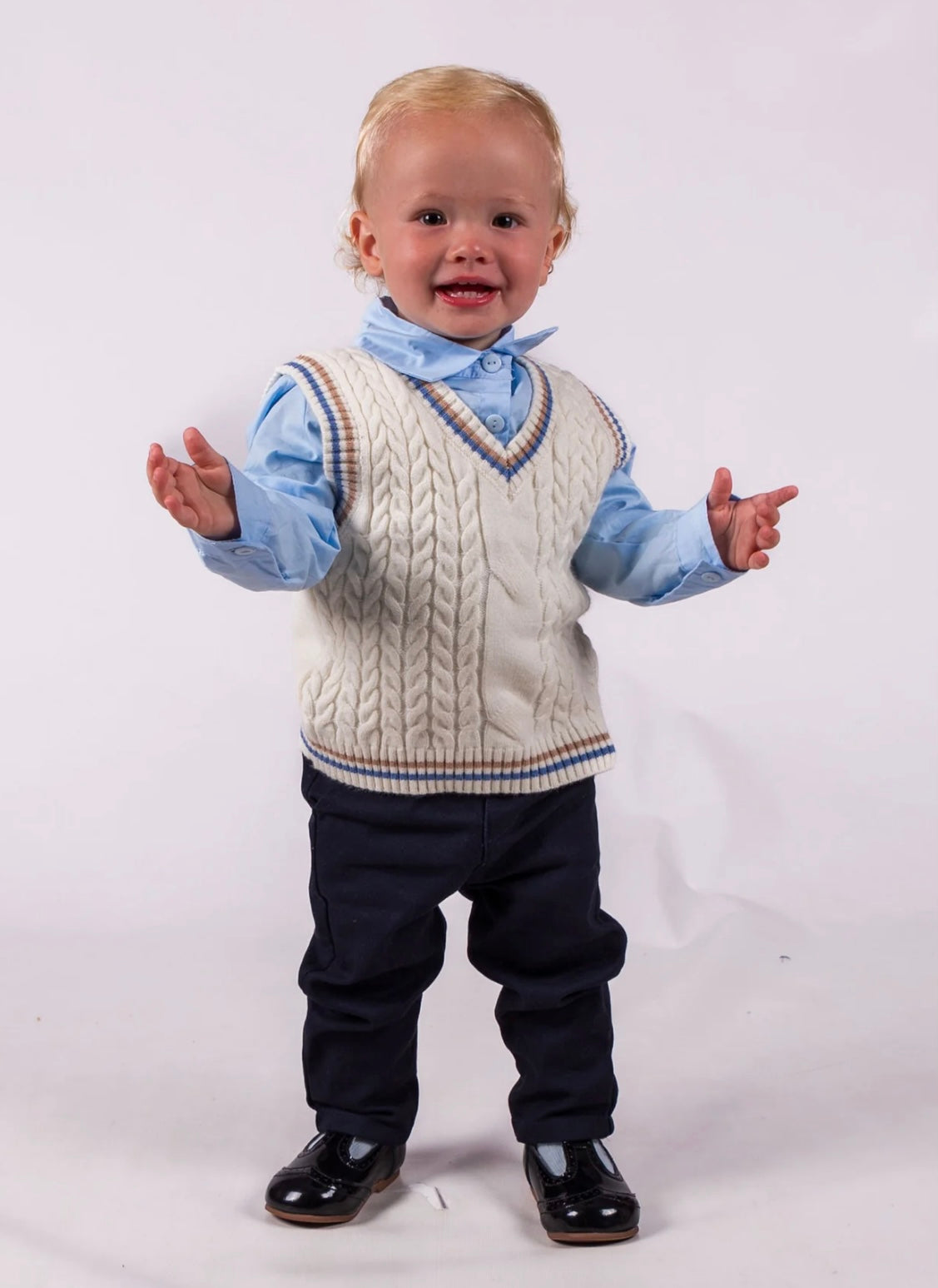 Baby wearing a cable knit vest over a shirt with rolled-up sleeves, black pants, and shiny shoes on a white background.
