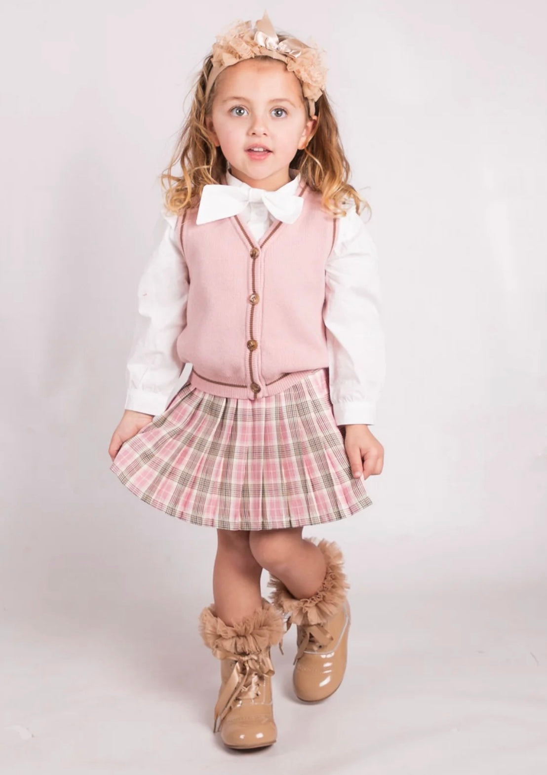 Young girl in a pink vest, plaid skirt, and white shirt with ruffled boots on a light background