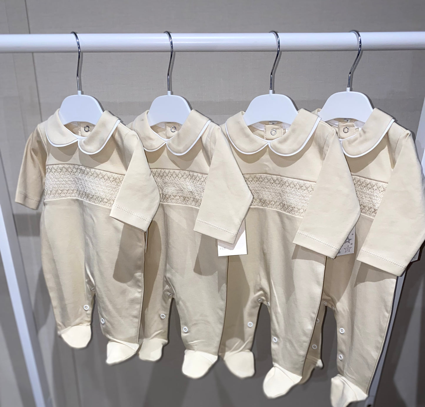 Four beige baby rompers with Peter Pan collars hanging on a rack against a gray background.