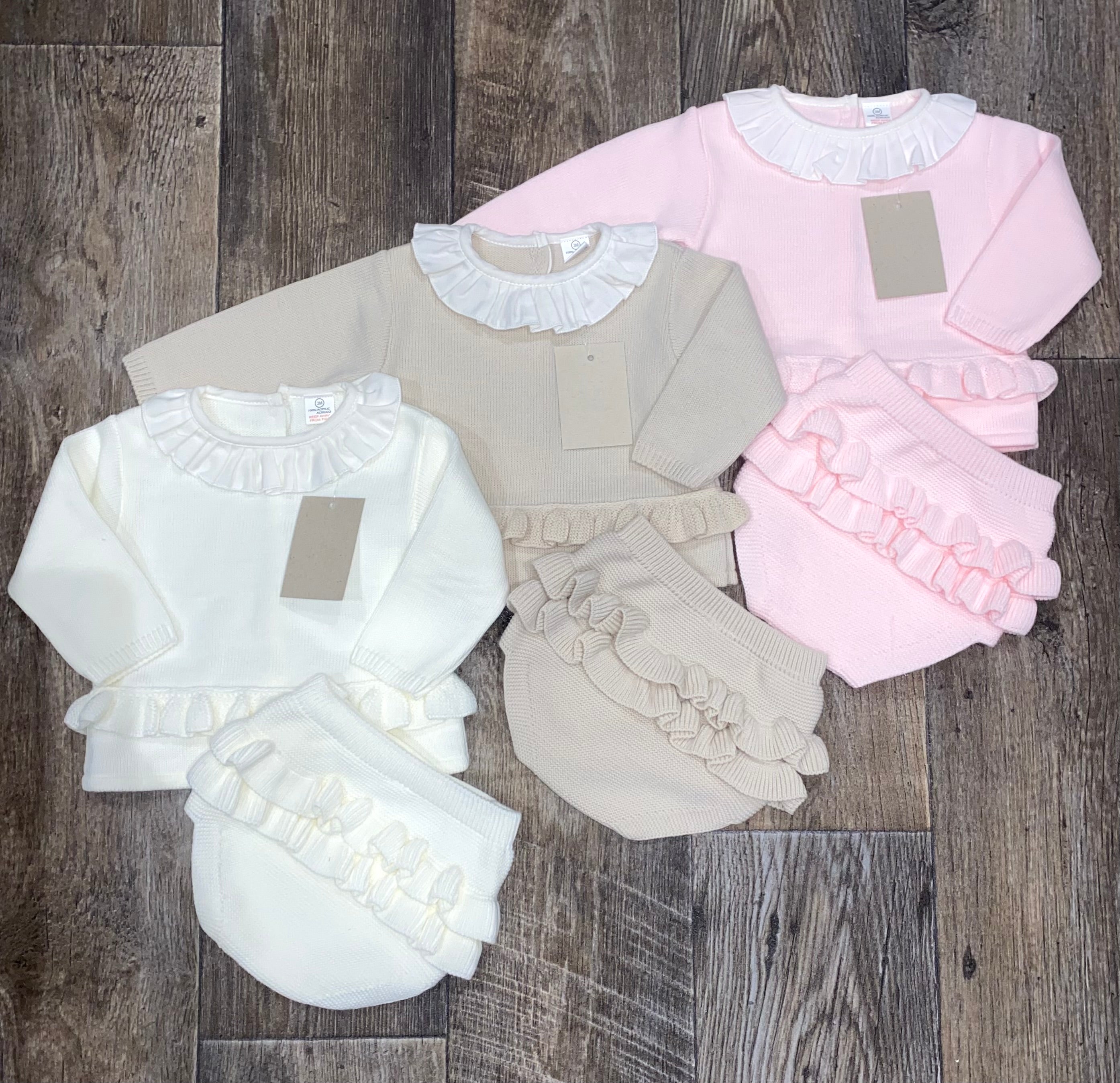 Three baby outfits in white, beige, and pink on a wooden surface