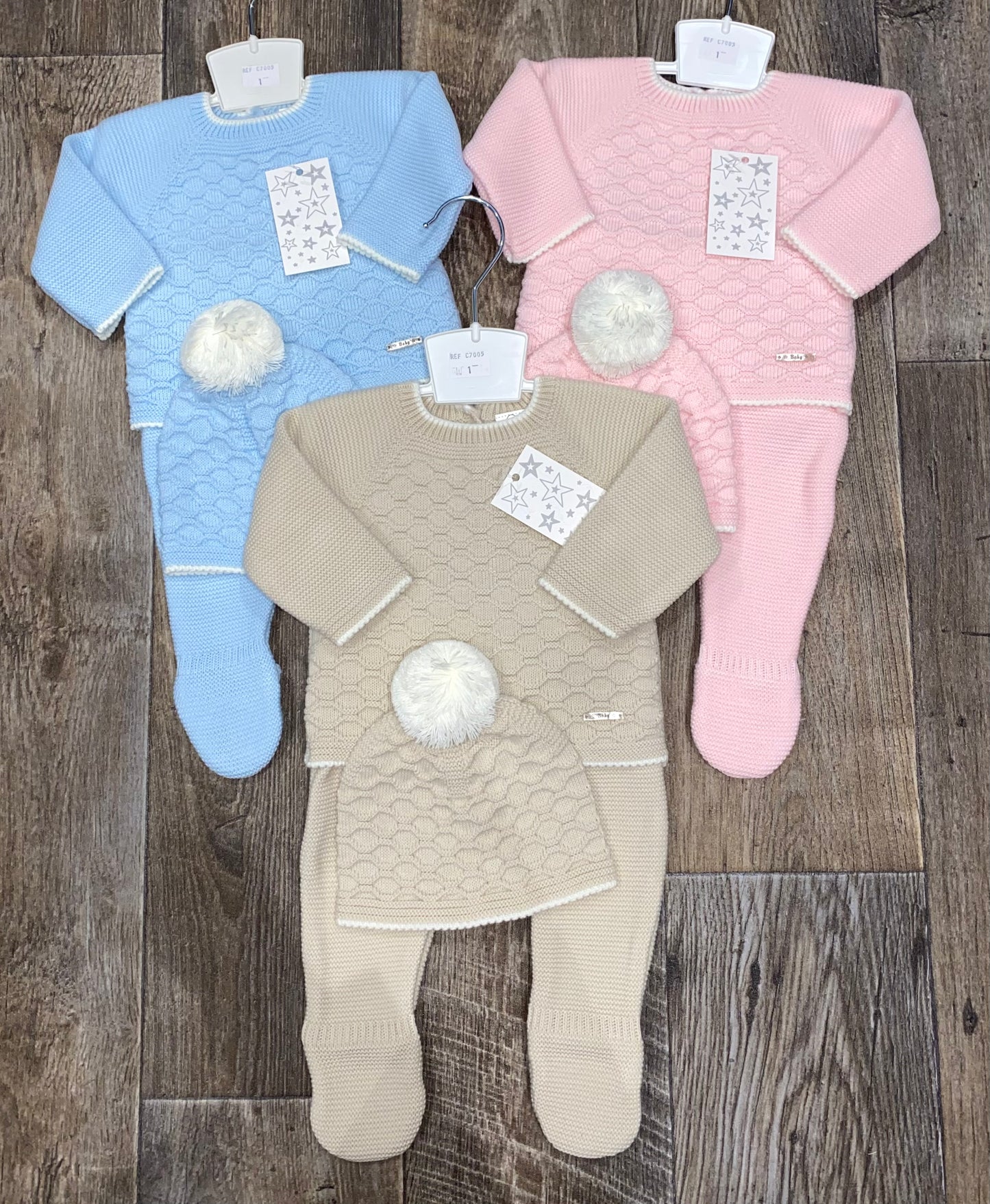 Three baby outfits in blue, beige, and pink on a wooden floor.
