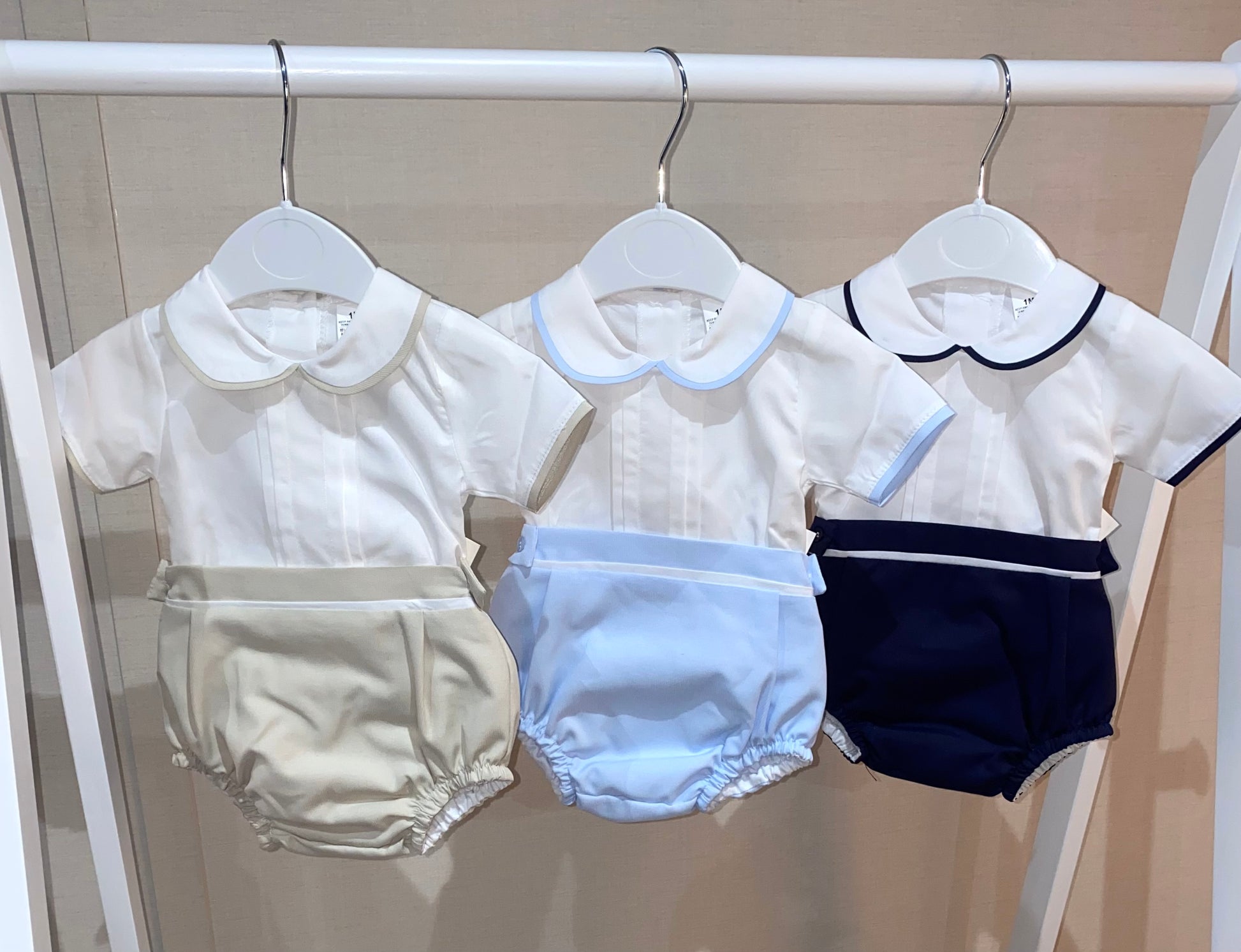 Three traditional baby rompers in cream, blue, and navy on hangers.
