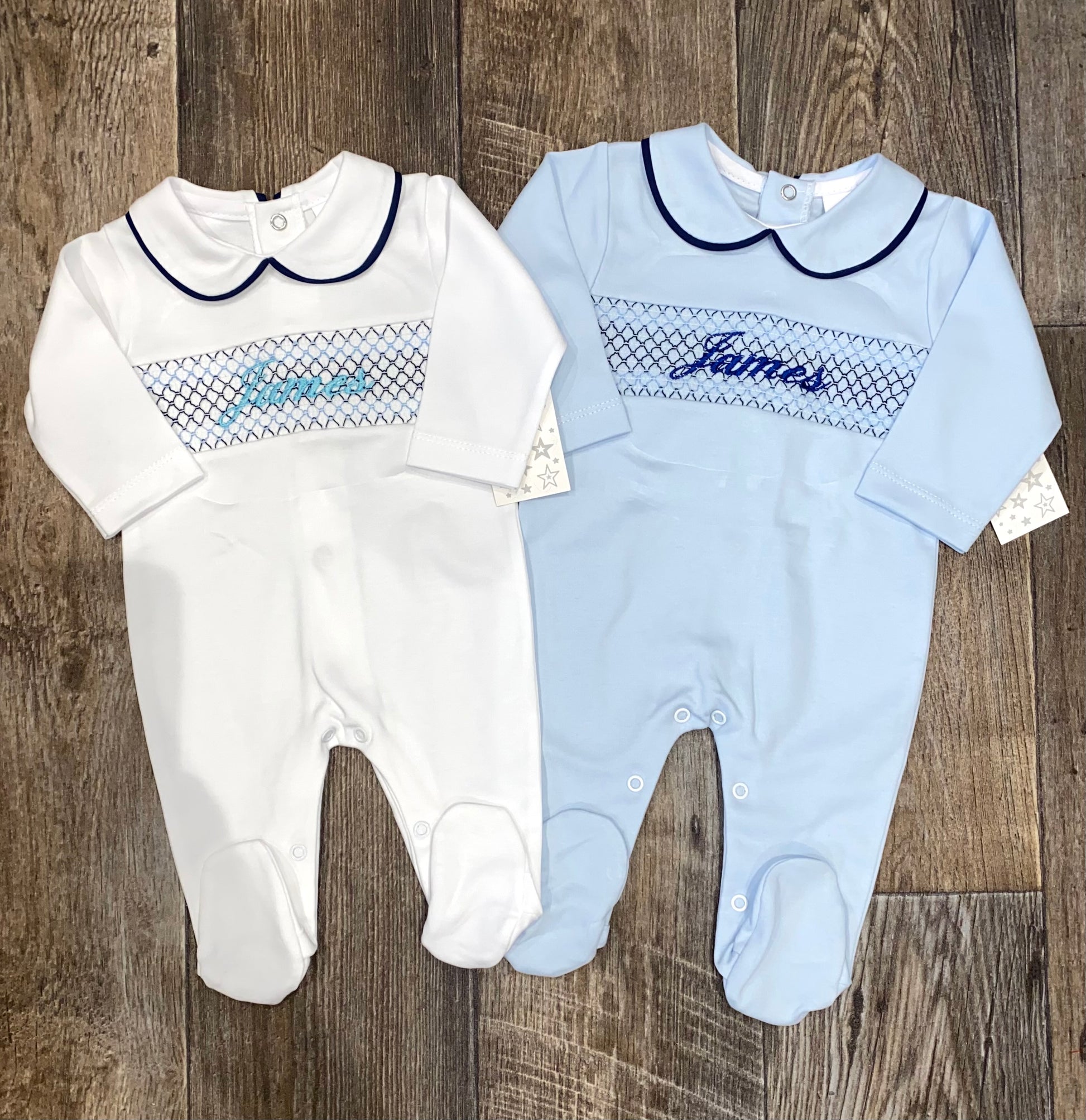Two baby rompers, one white and one blue, with embroidered names on a wooden floor.