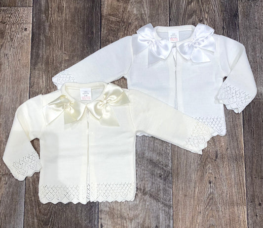 Two white baby outfits with lace details and bows on a wooden surface