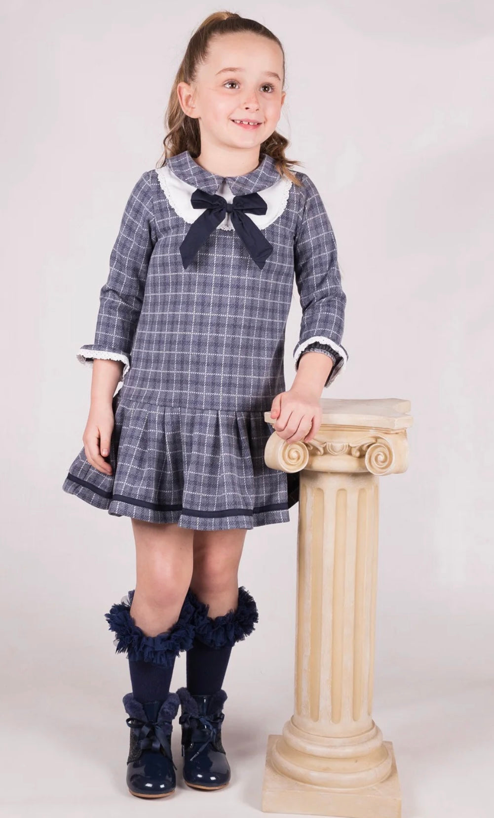 Young girl in a plaid dress with a large bow standing next to a decorative column on a white background
