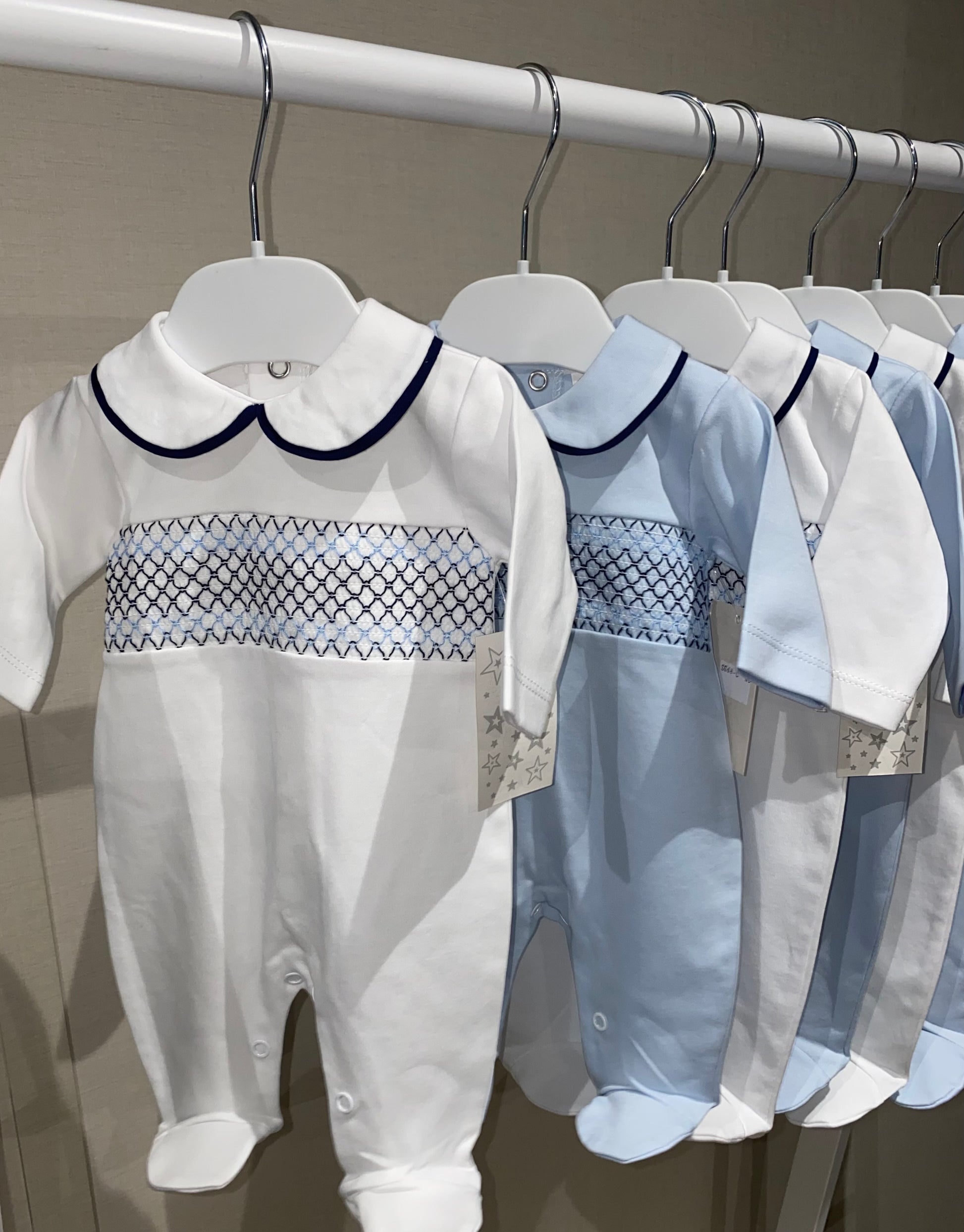 White and blue smocked baby rompers hanging on a rack against a neutral background