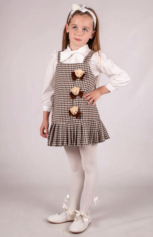 Young girl wearing a checkered pinafore dress with a white shirt and white shoes on a plain background