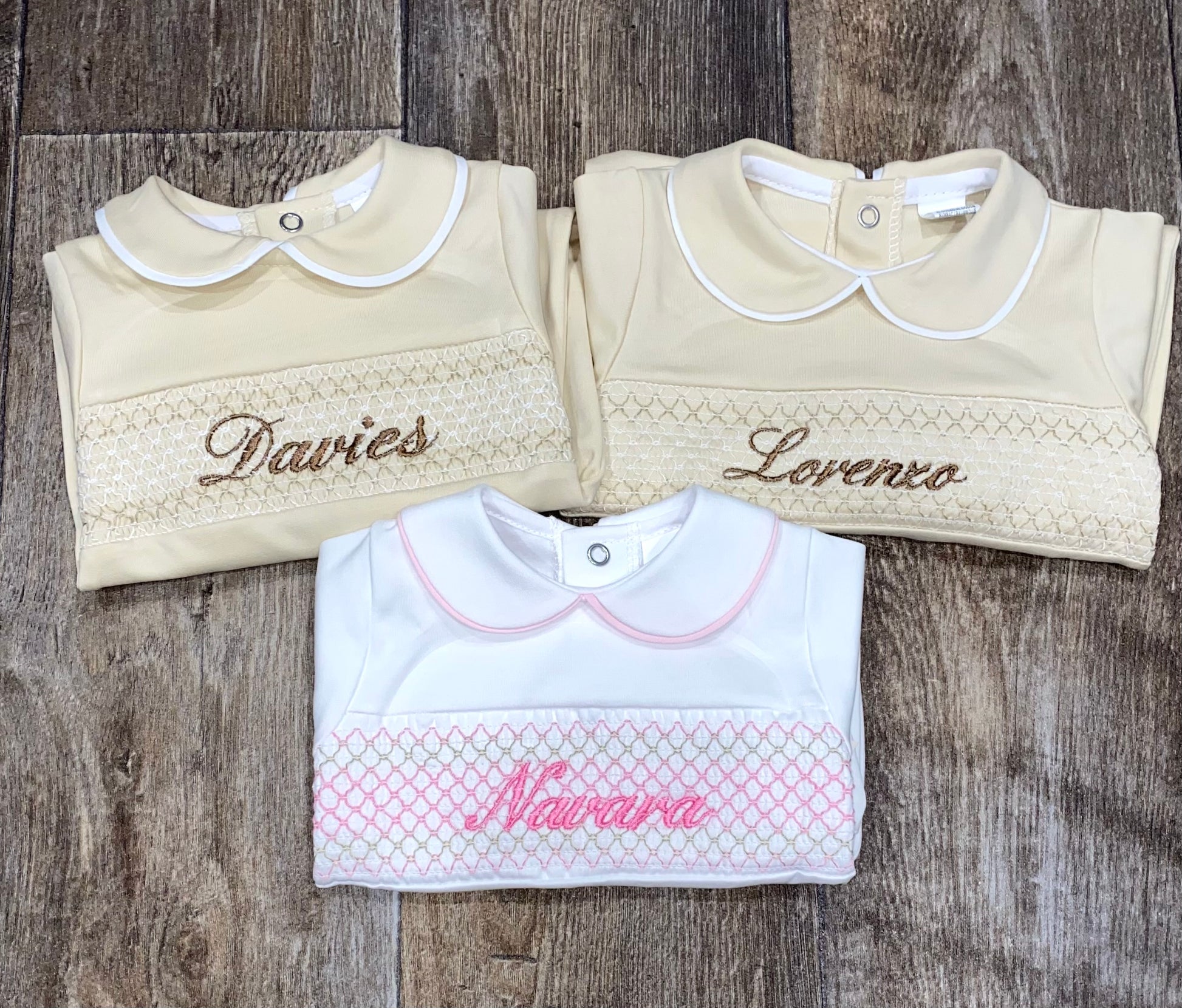 Three baby sleep suits with embroidered names on a wooden surface