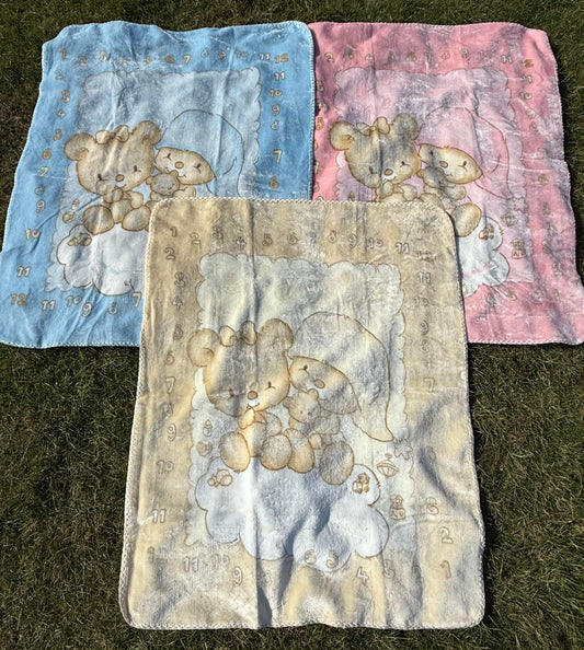Three baby blankets with teddy bear designs in pink, cream and blue