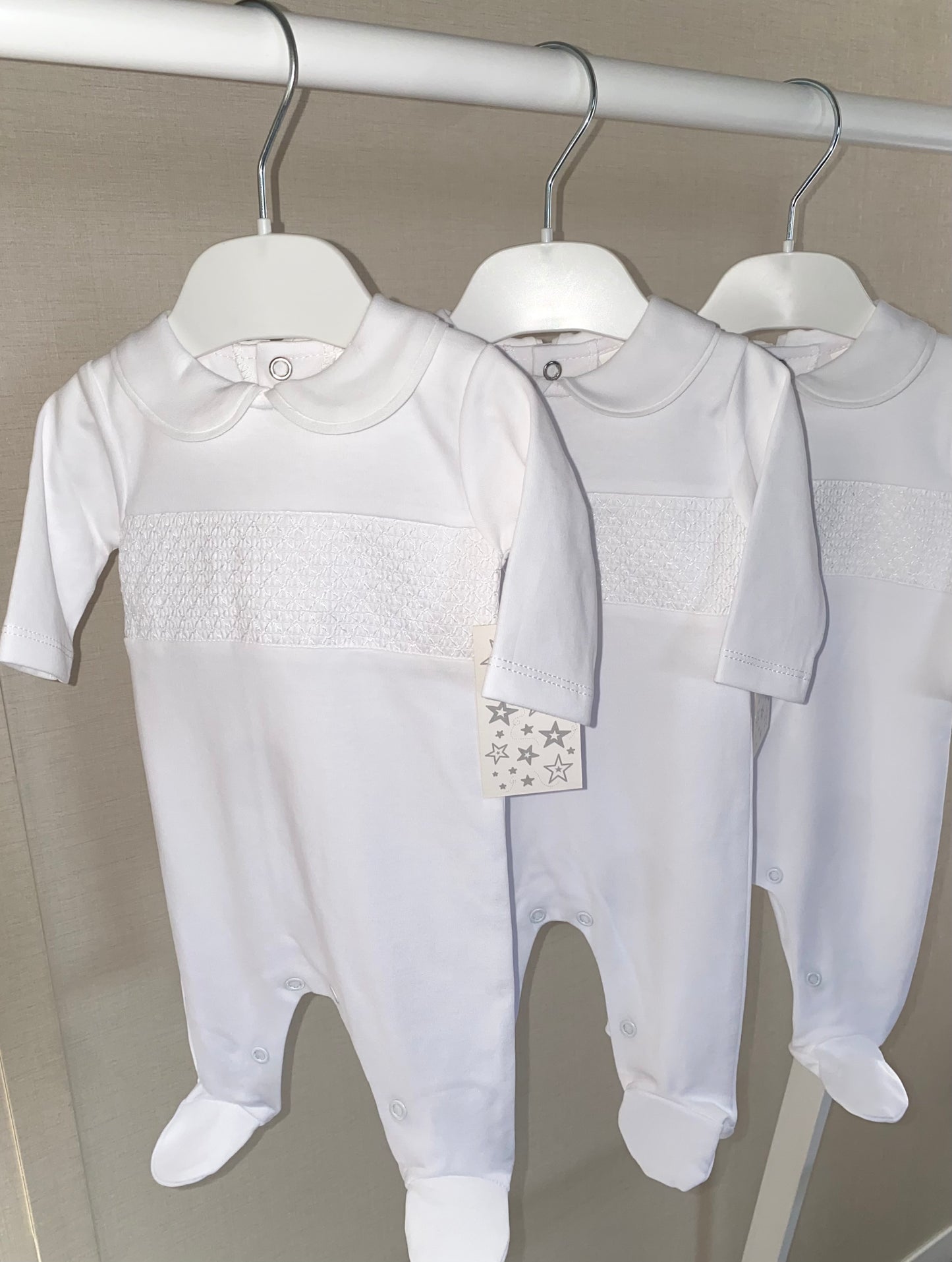 Three white baby rompers with smocking details on hangers against a neutral background