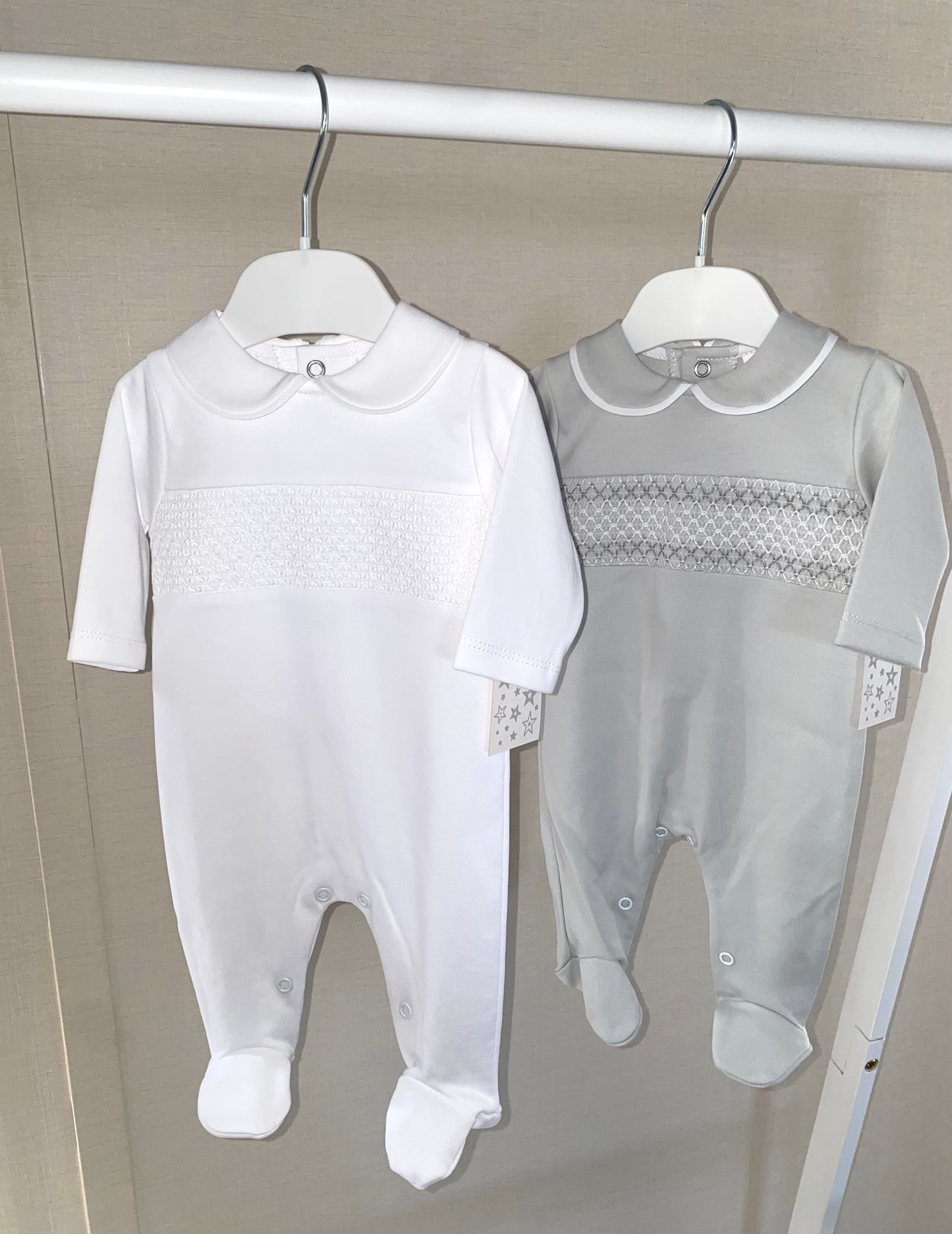 Two baby rompers, one white and one gray with smock details, hanging on a rack.