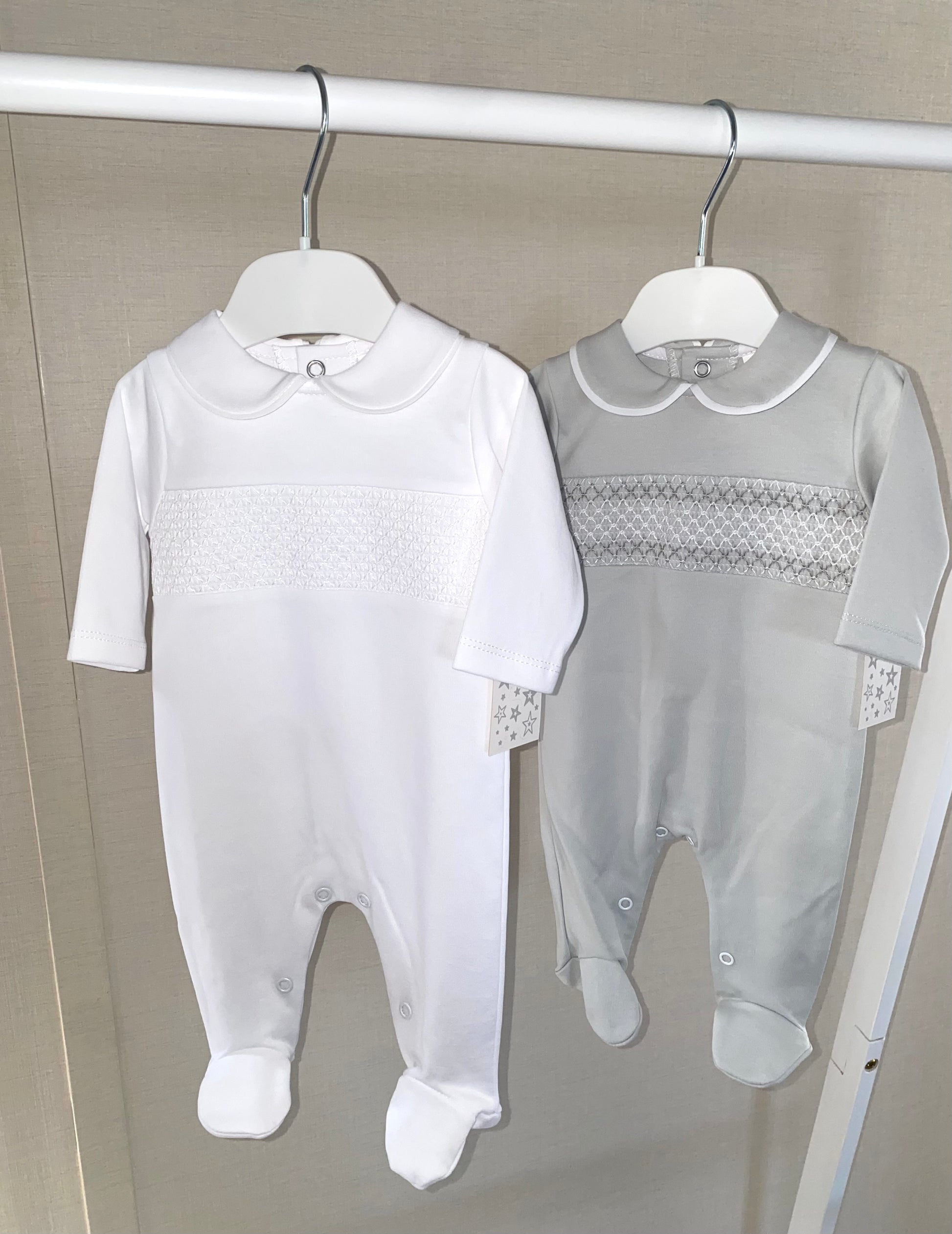 Two baby rompers, one white and one gray with smock details, hanging on a rack.
